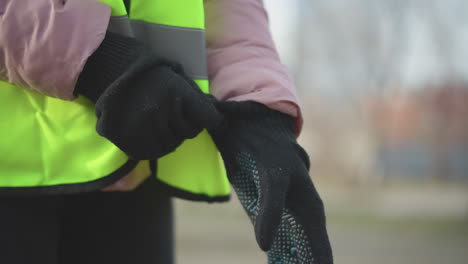 close-up of person wearing bright neon yellow high-visibility safety vest with reflective stripes and pink padded jacket putting on black knitted gloves outdoors, preparing for work