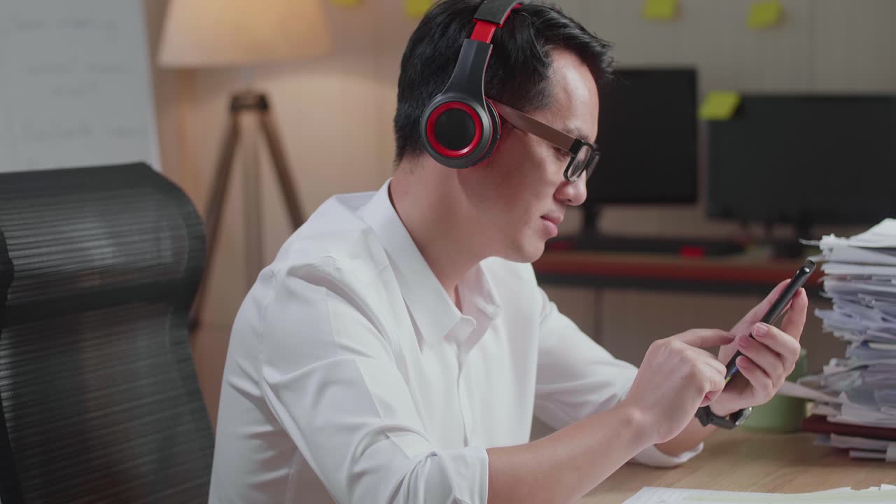 Close Up Of Asian Man With Headphones Listening To Music On Smartphone After Working With Documents At The Office