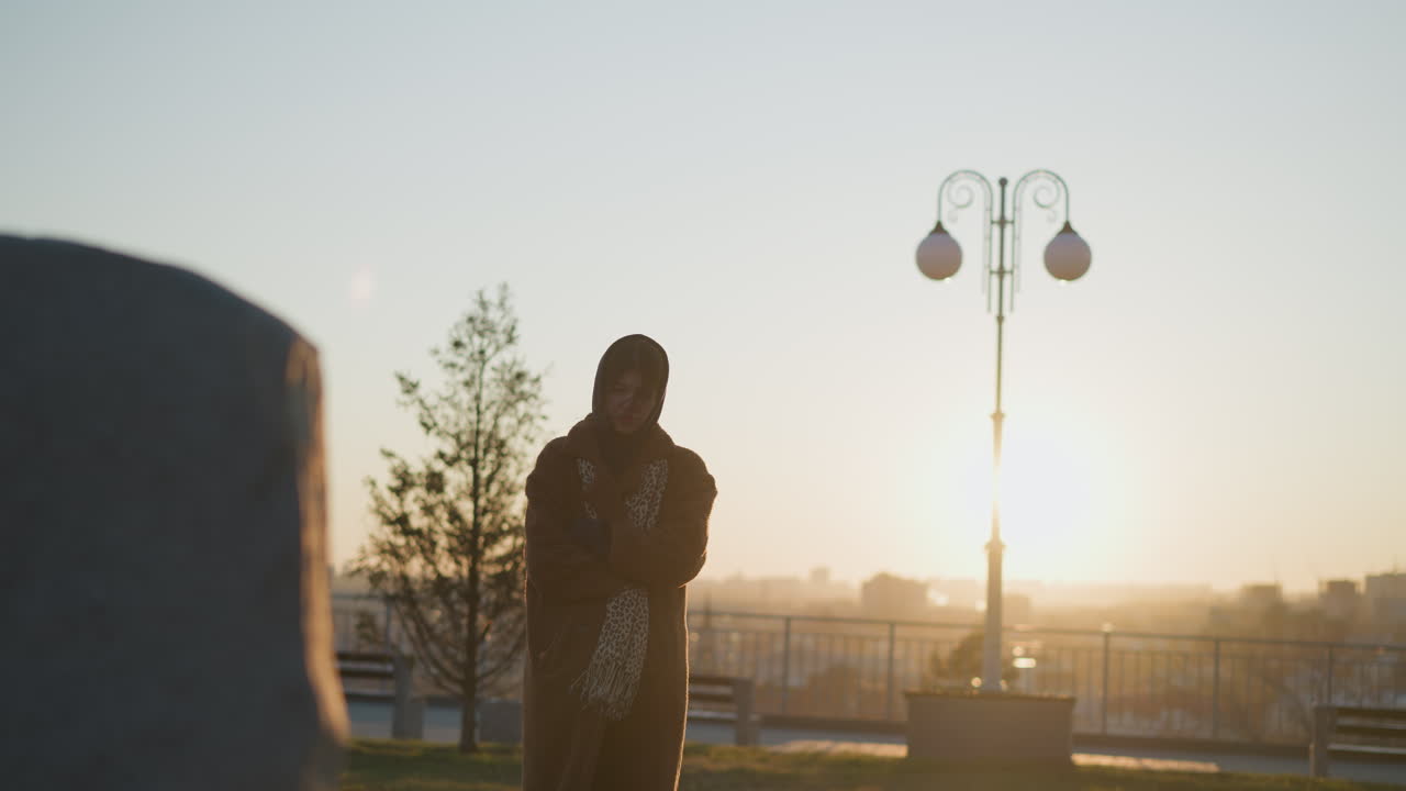 una niña llorona con las manos cruzadas en sus brazos, con un abrigo y una bufanda, de pie frente a un monumento de piedra en un parque durante la puesta del sol. su cabello cubre parcialmente la cara, reflejando su estado de ánimo triste