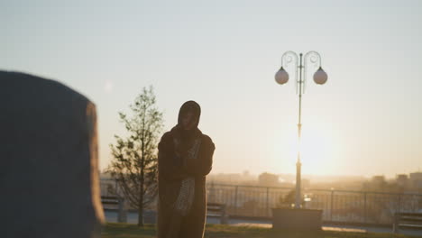 una niña llorona con las manos cruzadas en sus brazos, con un abrigo y una bufanda, de pie frente a un monumento de piedra en un parque durante la puesta del sol. su cabello cubre parcialmente la cara, reflejando su estado de ánimo triste