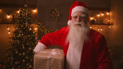 le père noël se tient dans le salon à côté d'un arbre de noël, puis prend une boîte avec un cadeau pour un enfant obéissant sous son aisselle pour un joyeux noël. regarde la caméra et sourit.