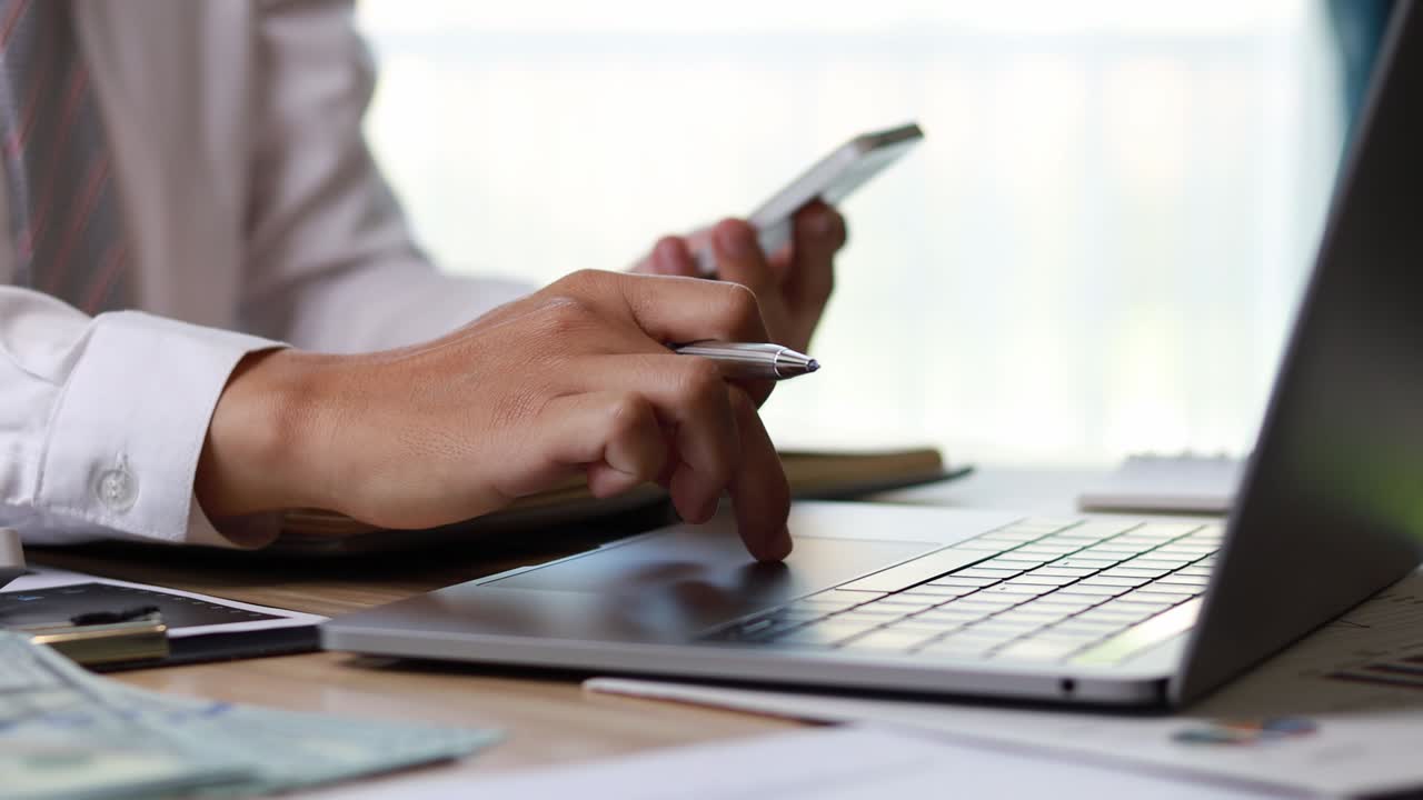 un hombre está frotando su dedo en el touchpad de una computadora portátil, con su mano izquierda sosteniendo un teléfono móvil, está navegando por la web para encontrar información. metraje 4k.