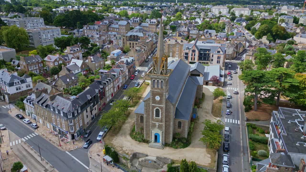 Notre Dame d'Emeraude church in Dinard, France, rises above traditional stone buildings and green trees, its tall steeple dominating the sunny coastal town skyline, drone orbiting shot