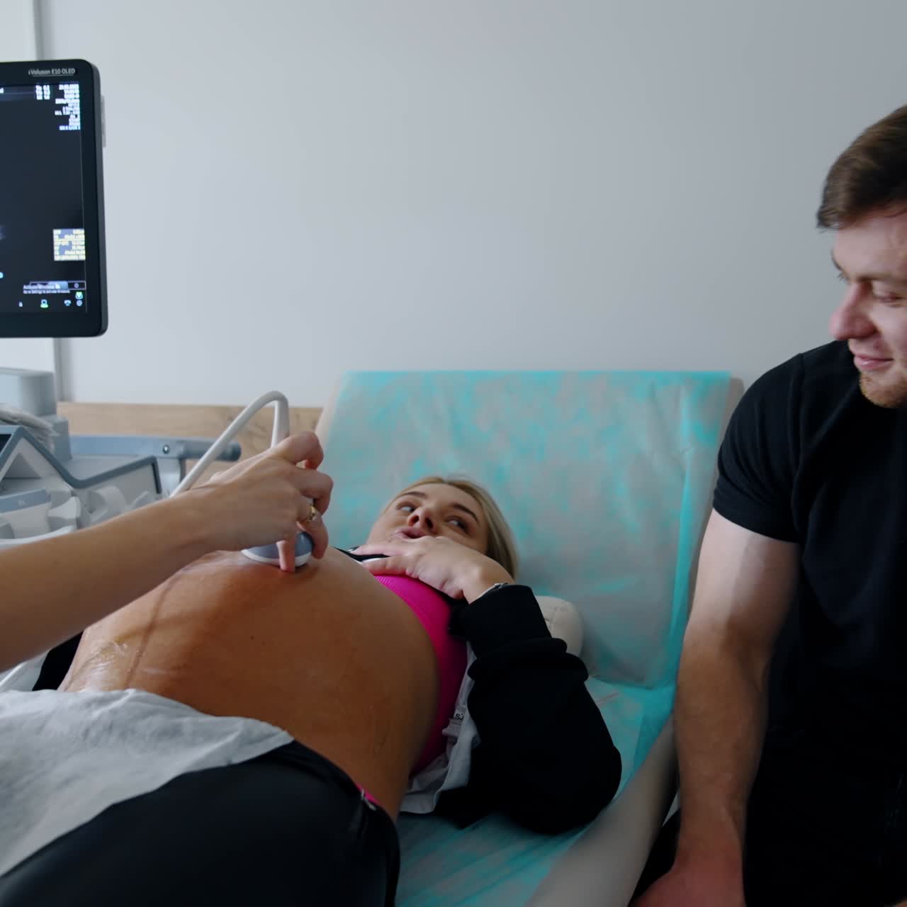 Happy future parents are having ultrasound examination of a baby. Lady lies on couch and examined by the doctor and her husband sits beside