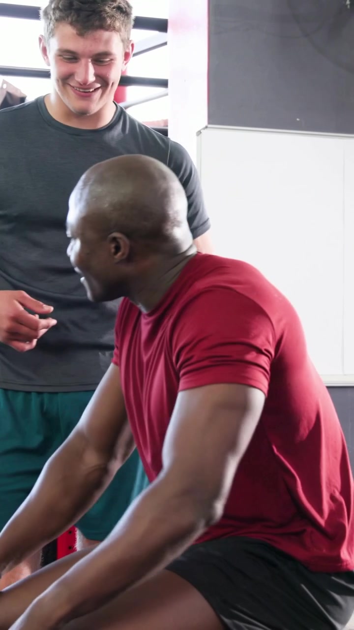 Vertical video: Stretching muscular man in red shirt massaging neck on plyo box at gym, taking tape