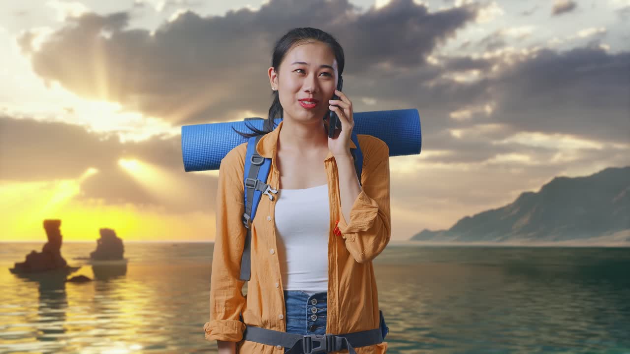 Nữ du khách đi bộ đường dài người châu Á đeo ba lô leo núi mỉm cười và nói chuyện trên điện thoại thông minh khi đứng trên bãi biển lúc hoàng hôn