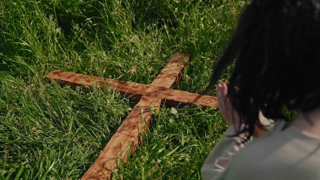 Over shoulder view of Jesus Christ looking down at wooden cross lying on green grass