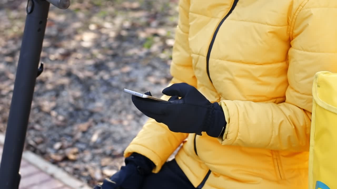 Food delivery man with scooter in a park sitting on a bench and using his smartphone. Yellow backpack and jacket. Winter
