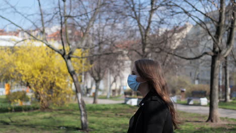 mujer joven con máscara protectora durante la pandemia del virus covid-19 caminando en un parque público en el soleado día de otoño, cámara lenta