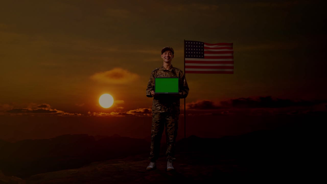 cuerpo lleno de hombre asiático soldado sonriendo y mostrando pantalla verde portátil a la cámara mientras está de pie con la bandera de los estados unidos, hora de la puesta del sol
