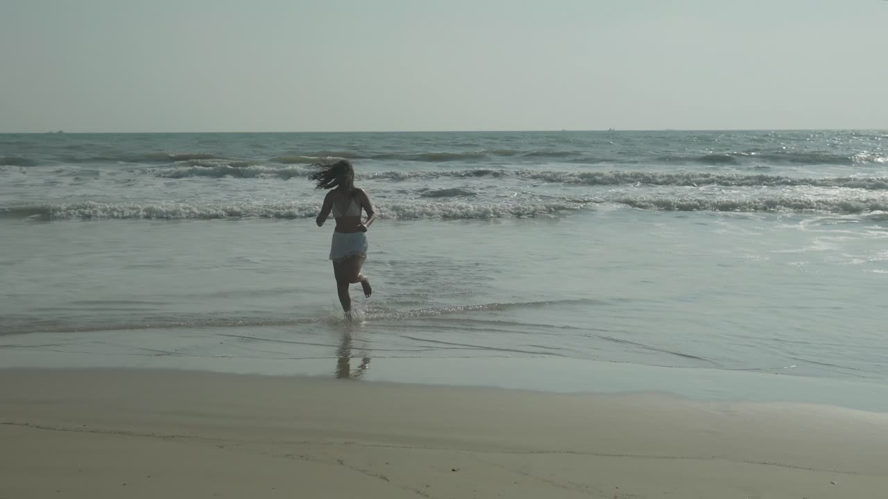 ⁇  비키니를 입은 어두운 머리카락의 여성이 바다에서 모래 해변으로 캐주얼하게 달리고, 따뜻한  ⁇ 빛 날