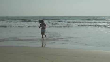 Mujer-Con-Cabello-Oscuro-En-Bikini-Blanco-Corriendo-Casualmente-Fuera-Del-Mar-Hacia-La-Playa-De-Arena,-En-Un-Cálido-Día-Soleado