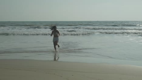 woman with dark hair in white bikini running casually out of sea onto sandy beach, on warm sunny day