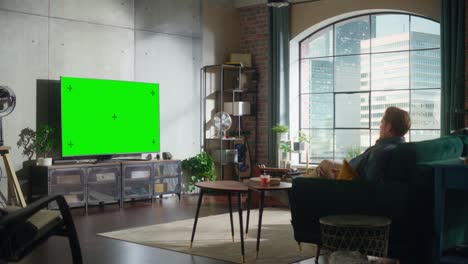 joven hombre guapo pasando el tiempo en casa, sentado en un sofá y encendiendo la televisión con pantalla verde simulacro de exhibición en un elegante apartamento loft. hombre transmisión de la película en casa.