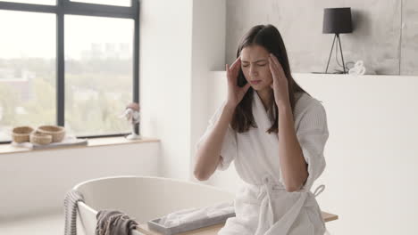 Suffering-Woman-In-Robe-Having-Headache-While-Sitting-On-Bathtub-In-A-Modern-Bathroom