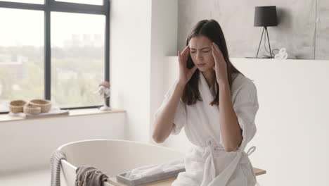 suffering woman in robe having headache while sitting on bathtub in a modern bathroom