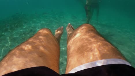 underwater subjective perspective of man legs floating in clear transparent sea water beneath surface with people in background