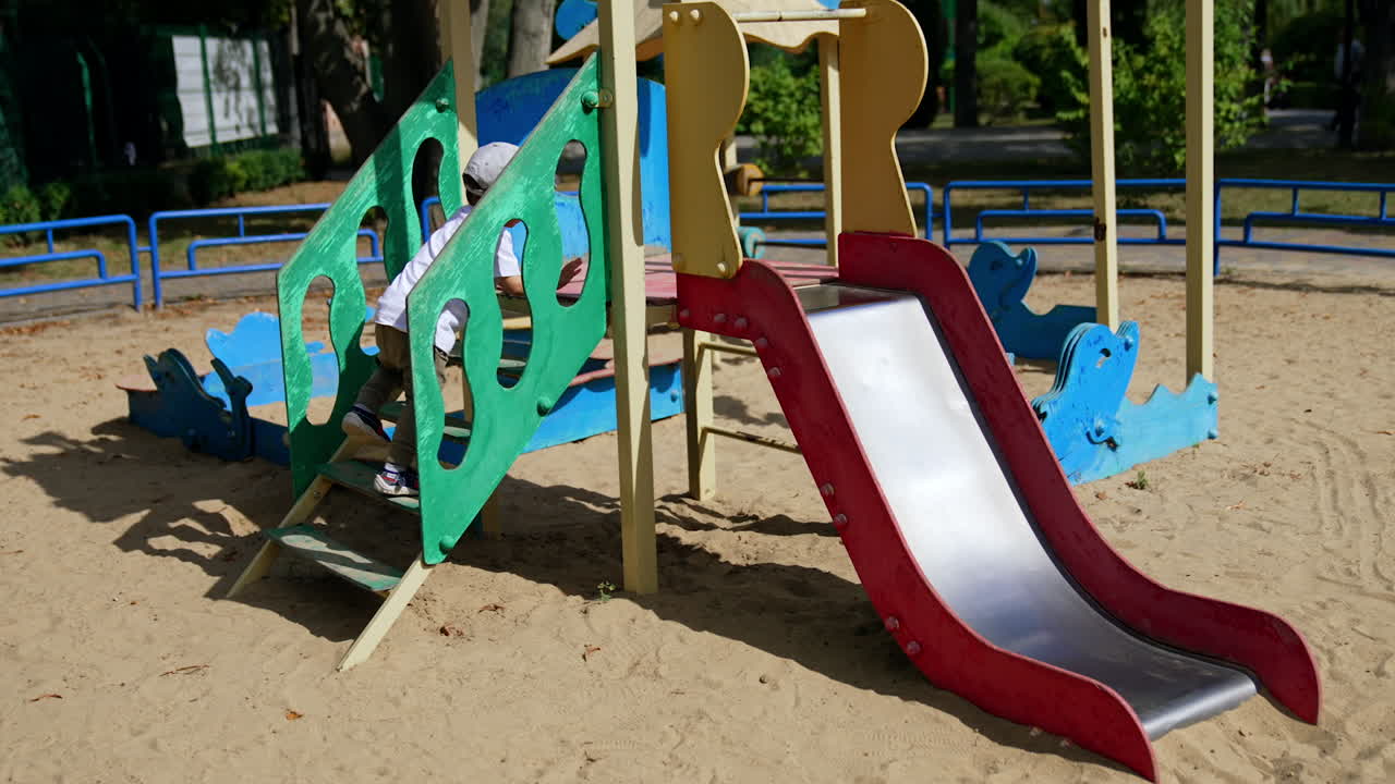 Male toddler goes up slowly by the slide on the playground. Baby sits down on the slide and taps his feet by it.