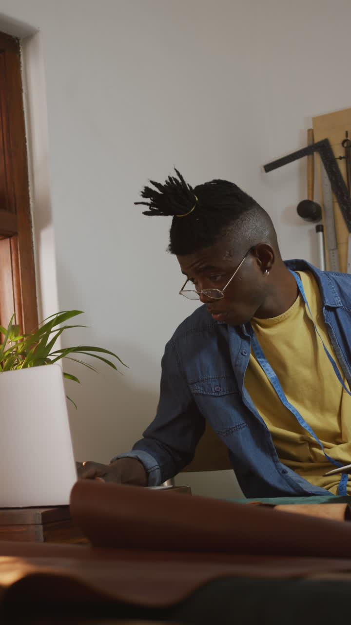 Vertical video of african american leather craftsman with dreadlocks using laptop in workshop