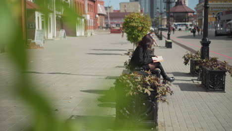 primer plano de hojas verdes balanceándose en el viento con una mujer sentada con la pierna cruzada, leyendo un libro, un coche lejano pasa con un fondo borroso con paisajes urbanos y plantas en maceta