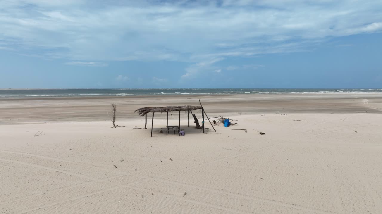 Shot of a makeshift shelter made of wooden poles and thatched roof in a vast, sandy beach with Parnaiba river at background in Brazil.