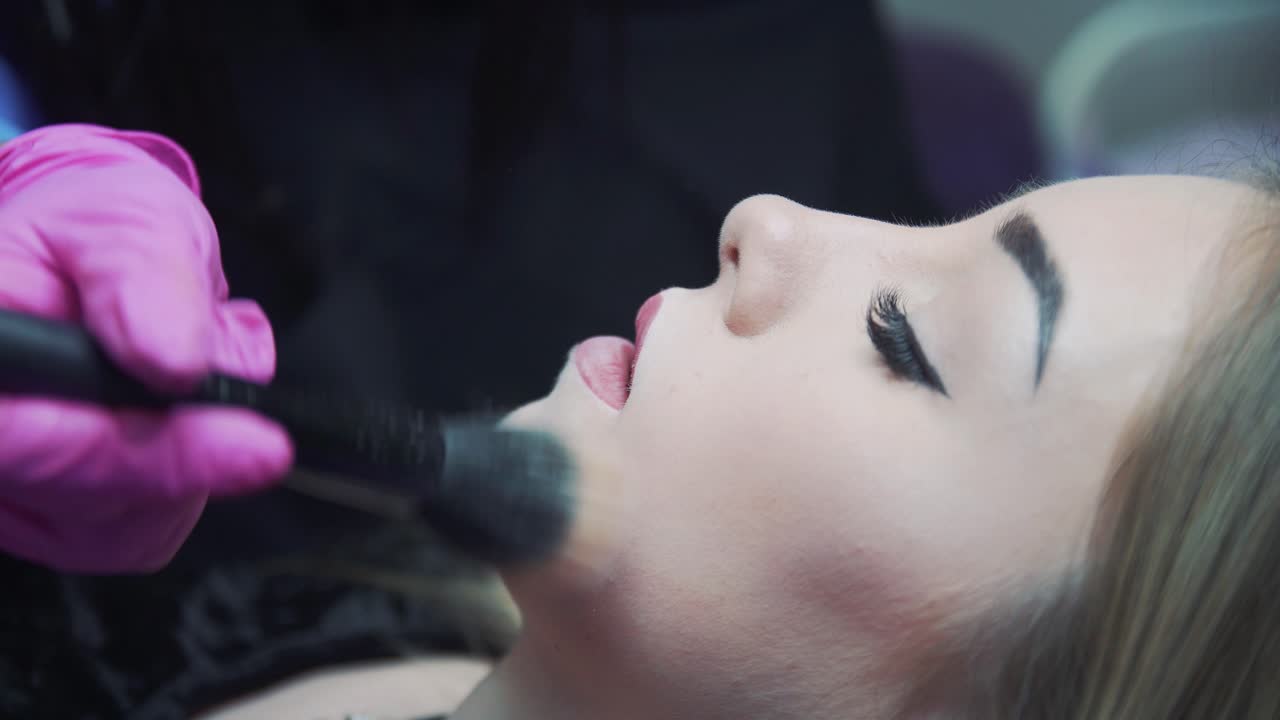 Makeup for a girl in a beauty salon. The cosmetologist with a brush applies makeup powder on the face