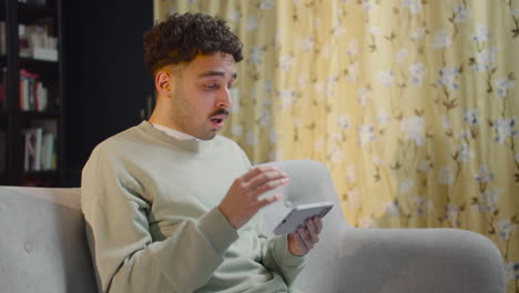 young man sitting on sofa and watching some shocking movie scene on smartphone