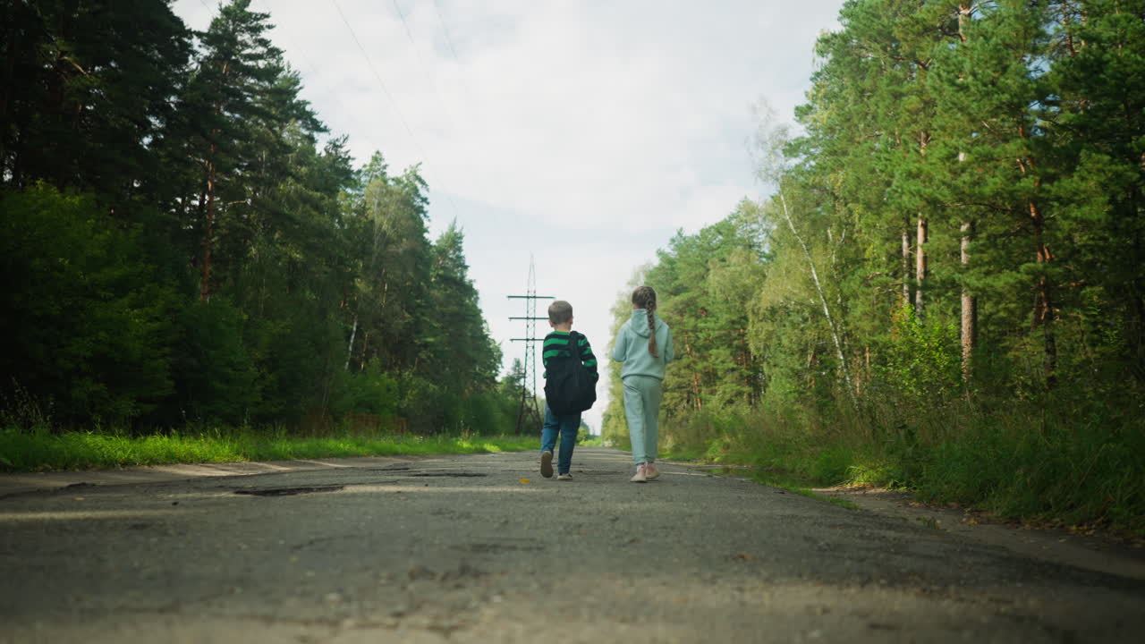 Rear view of two children walking side by side along tarred road in forested area with power cables stretching into distance under partly cloudy sky