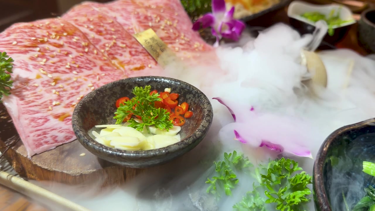 Primo piano di manzo wagyu crudo marmorizzato servito con aglio, peperoncino e ghiaccio secco fumante.