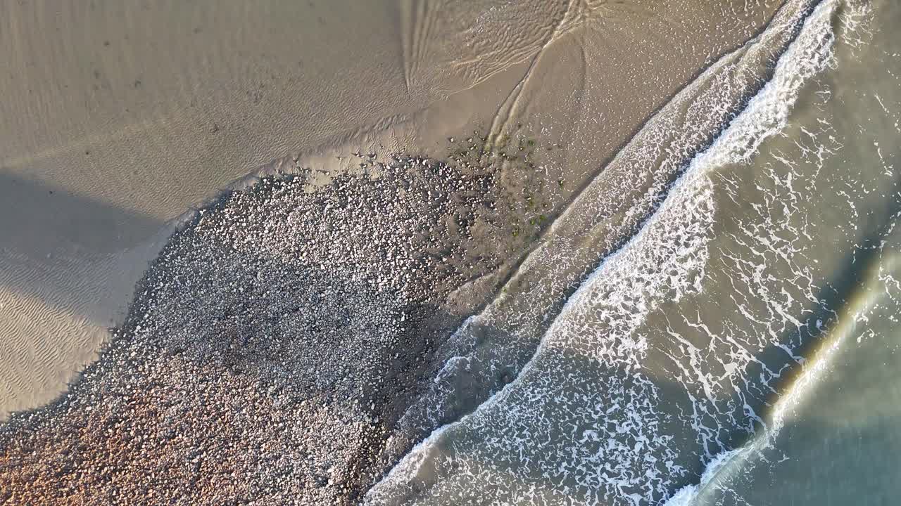 Drone captures waves gently crashing on a rocky beach, creating dynamic patterns and shadows in natural light