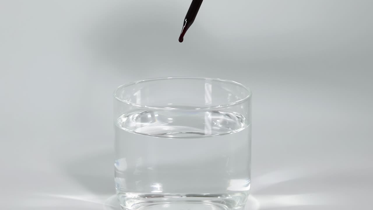 A single ink droplet falls into a glass of clear water, creating a visual effect.