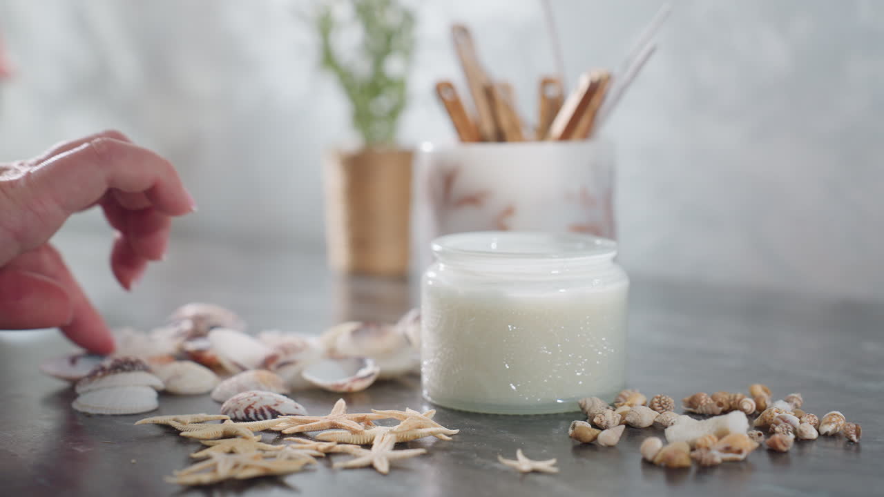 Close up of woman hand gently arranging decorative seashells, tiny starfish, and pebbles on smooth marble table beside white candle jar and holder