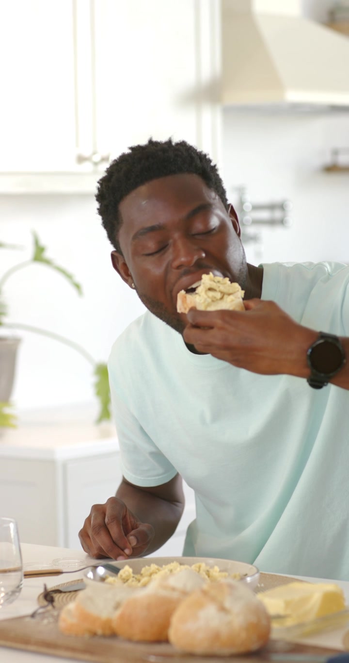 Vertical video: African American man picking up roll from plate in kitchen, biting spread to eat