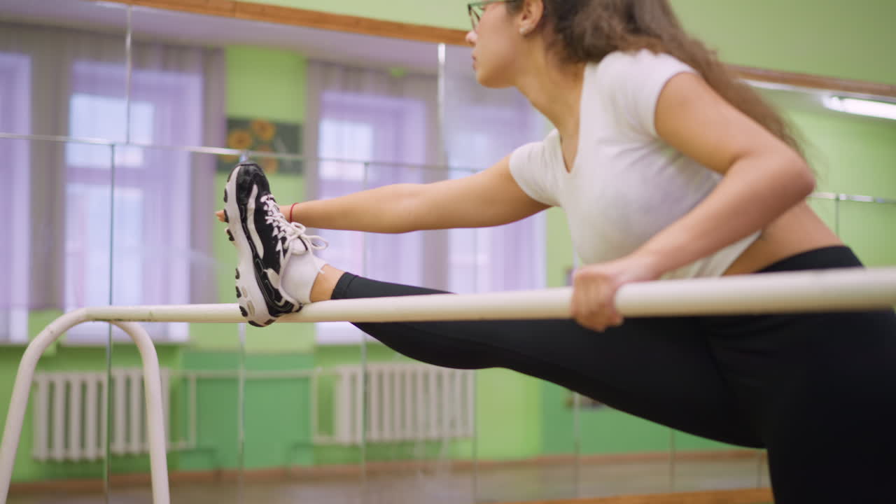 Young girl in white top and black leggings stretching leg on bar indoors, sneakers and socks visible, arm extended for balance, workout focused on flexibility, strength, and fitness training