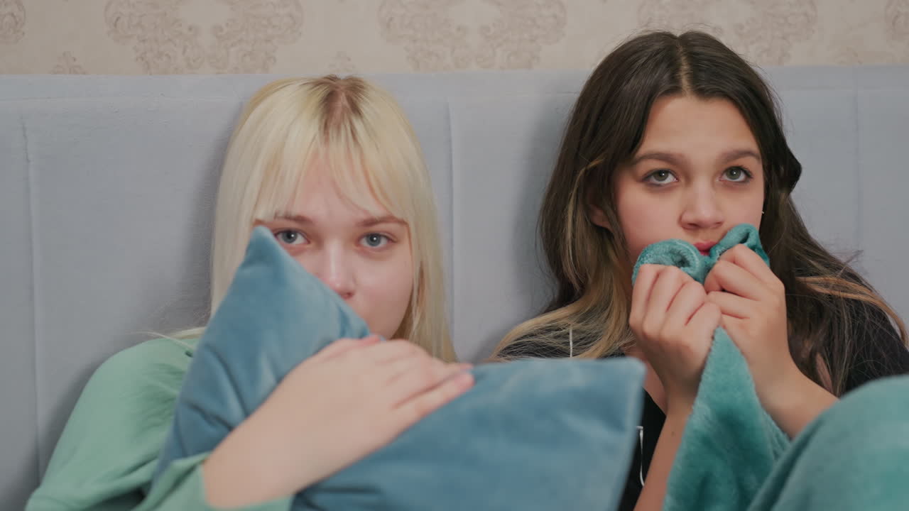 two teenage girls sitting on bed looking frightened while watching horror movie, one clutching soft pillow and other biting blanket, both with wide eyes and tense expressions