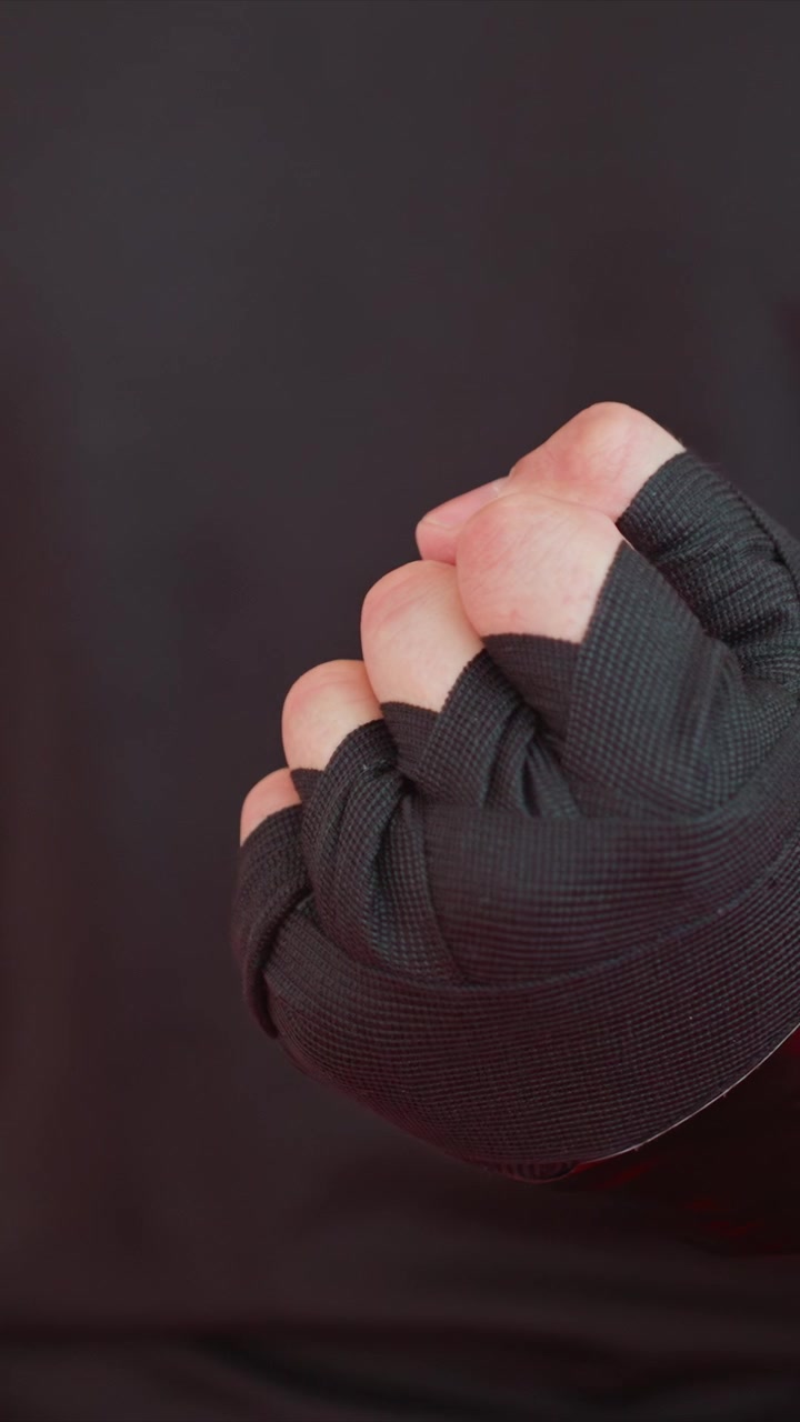 Close up of hands being wrapped with black hand wraps focusing. Hands move slightly as wrapping progresses. Dark background emphasizes close up view of gloved fists