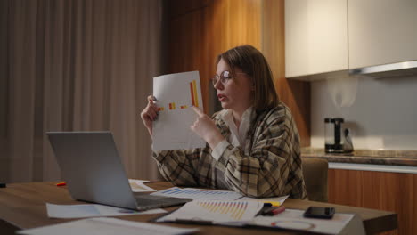 A-young-woman-with-glasses-at-home-shows-a-graph-in-a-laptop-camera-sitting-at-home-in-the-kitchen-conducting-an-online-report.