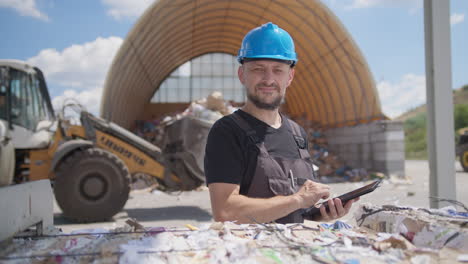 Gerente-De-La-Planta-De-Reciclaje-De-Papel-Mira-La-Cámara,-El-Cargador-De-Ruedas-Detrás-Mueve-La-Basura