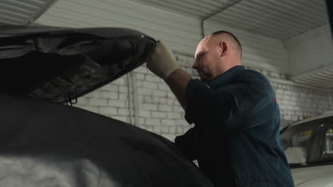 ingeniero de automóviles en uniforme azul habilmente utiliza una cuña para mantener abierto el capó de un coche negro en un taller mecánico bien iluminado, inspeccionando cuidadosamente los componentes del motor para el mantenimiento y la reparación