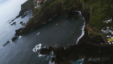 aerial rolling drone view of seixal village on a dramatic volcanic coast, with natural lava rock swimming pools and a dark sand beach, madeira, portugal