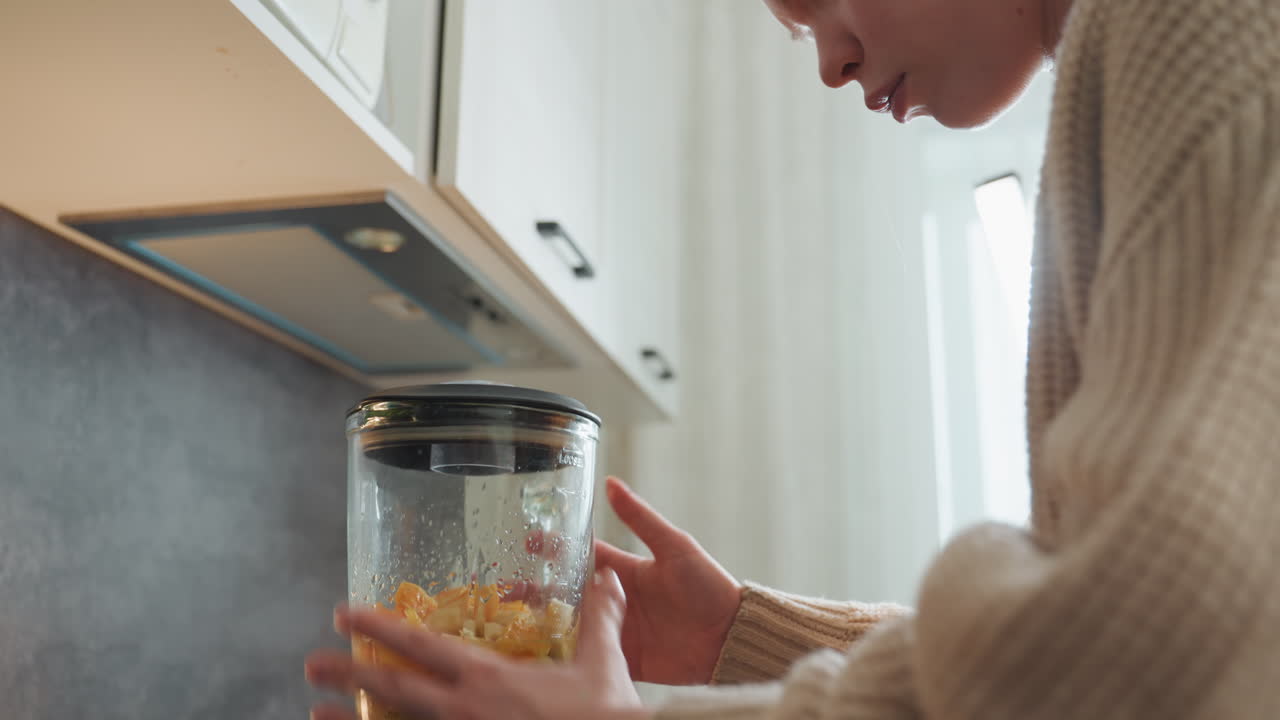 Primo piano di una donna incinta che controlla il coperchio e le impostazioni del frullatore in una cucina compatta, assicurandosi una chiusura sicura prima di frullare, misura gli ingredienti con espressione concentrata, genitore in attesa con maglione, luce soffusa.