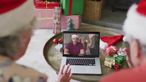 Senior-caucasian-couple-with-santa-hats-using-laptop-for-christmas-video-call-with-couple-on-screen
