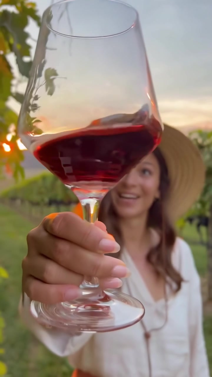 Giovane donna che tiene un bicchiere di vino rosso in un vigneto al tramonto.