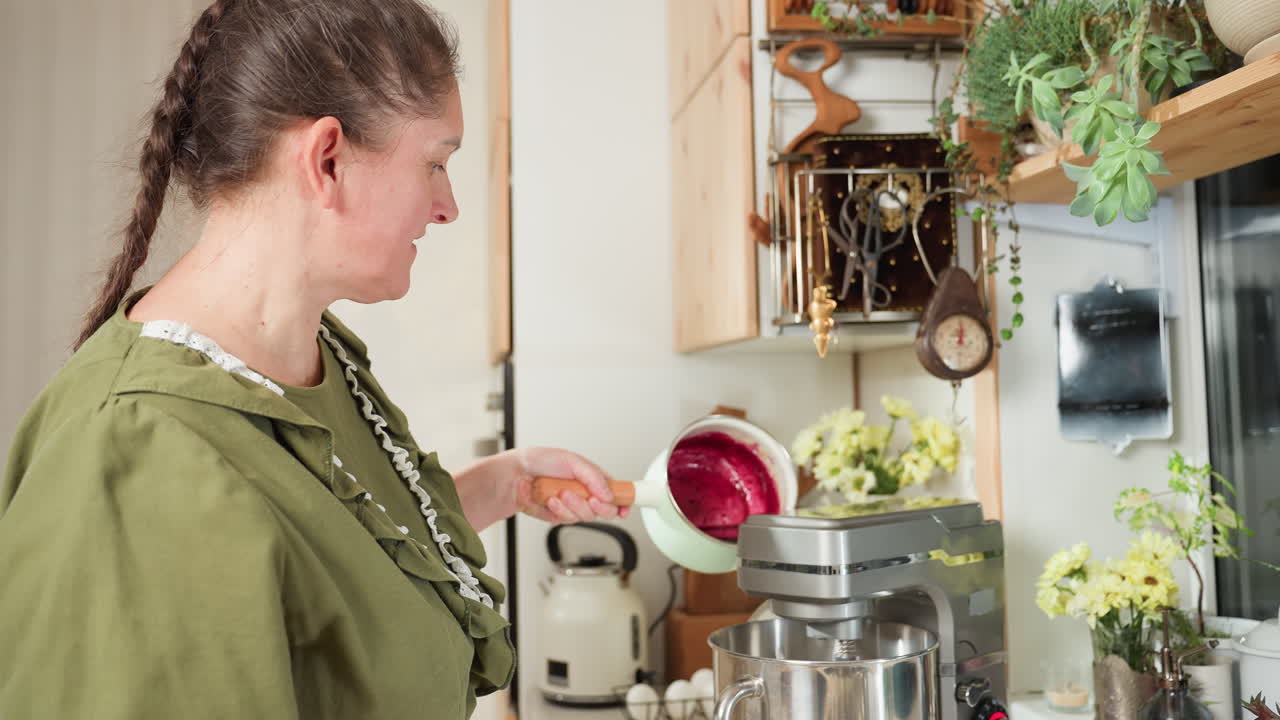 Woman in green outfit pouring blended red currant mixture from saucepan into electric stand mixer bowl in cozy kitchen filled with natural light, rustic elements, flowers, and hanging utensils