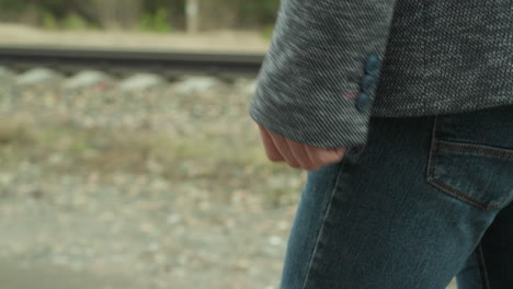 vista en primer plano de una persona con pantalones vaqueros azules y una chaqueta gris sosteniendo una pistola mientras camina por un camino de tierra, con un fondo de bosque borroso