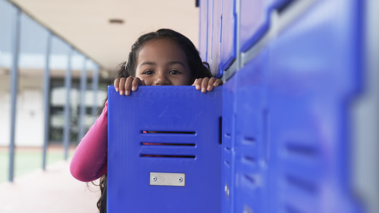 In a school hallway, a young biracial girl peeks from behind a blue locker