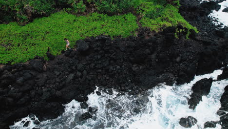 drone shot of adventurous young woman walking on sharp lava rocks over crashing waves