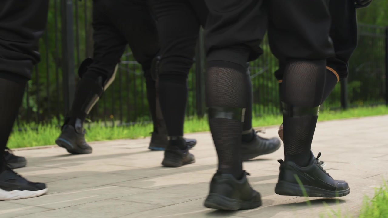 Close up of multiple people legs and feet in synchronized movement wearing black clothing with knee pads. Footwear includes various styles of athletic shoes on paved ground near green grass