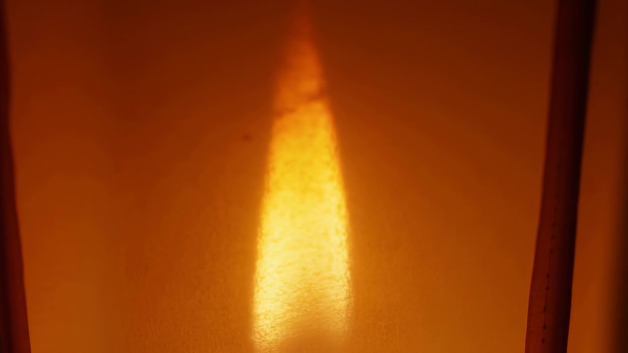 Un brillante bagliore ambrato verticale si intensifica e si allunga da un debole riflesso in un'alta colonna luminosa e ristretta contro una superficie opaca arancione calda; un punto di vista ravvicinato dal basso enfatizza la profondità di.