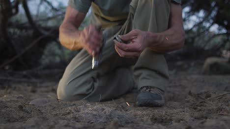 caucasian male survivalist using knife and flint to spark fire with tinder at camp in wilderness